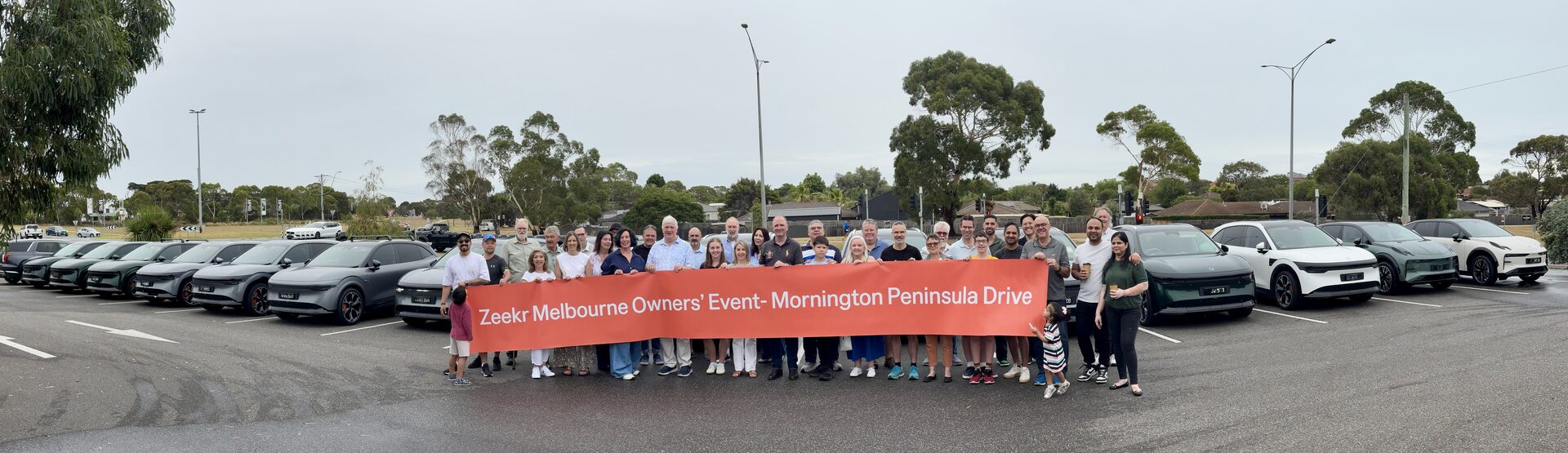 ZEEKR owners and families standing behind a Mornington Peninsula Drive banner, with a long line of ZEEKR vehicles behind them.